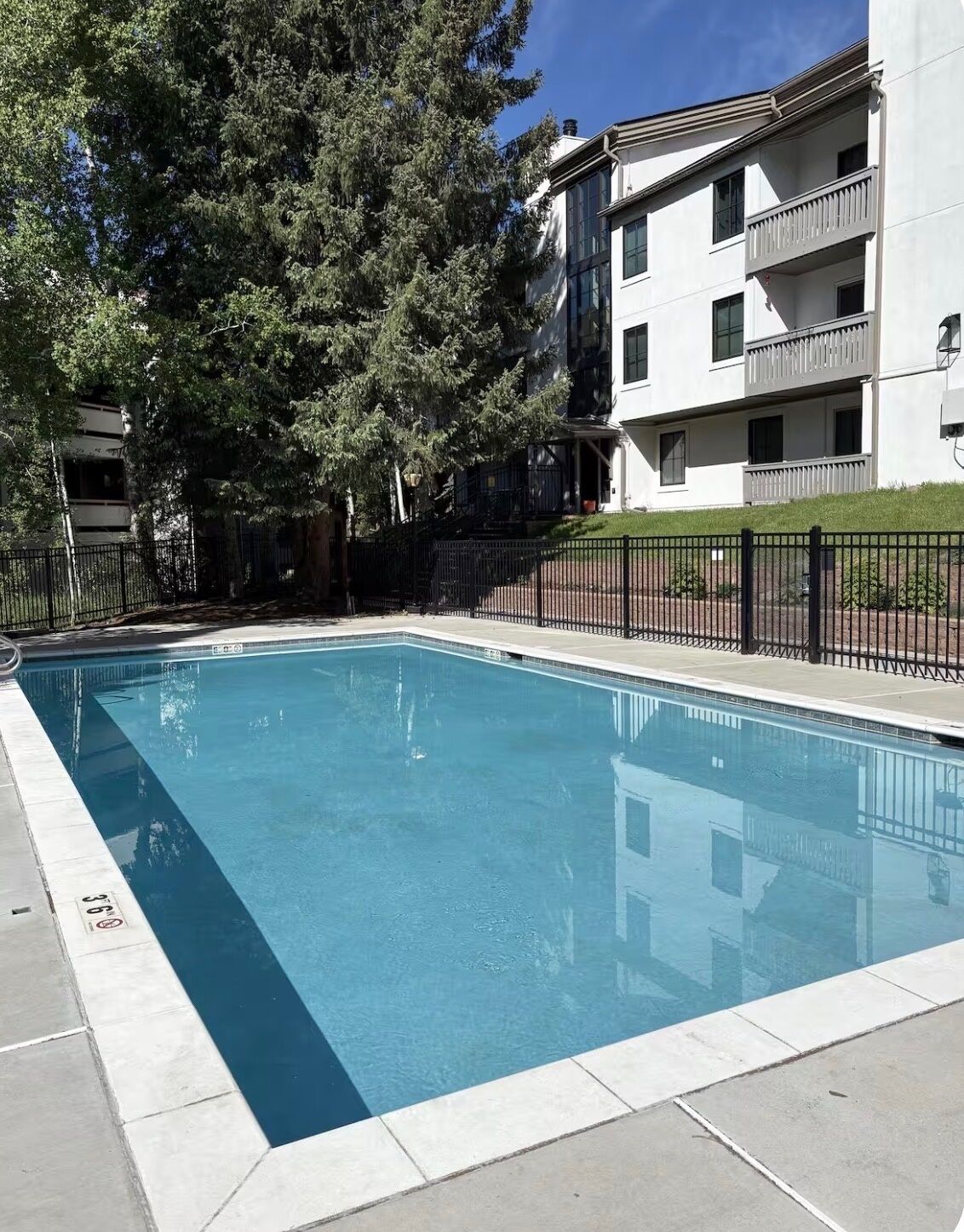 A rectangular swimming pool with a tiled edge, surrounded by a black metal fence, and adjacent to a multi-story building with balconies.