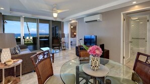 A modern living room with a glass dining table, comfortable seating, a flat-screen TV, and a view of the ocean. - Royal Mauian, #116^ 2 Bedroom Condo (Kihei)