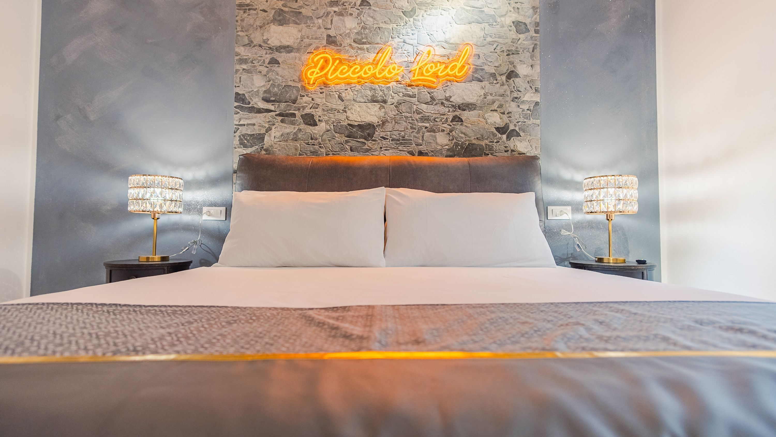Uma cama bem arrumada com uma parede de pedra ao fundo, duas luminárias de cabeceira e um letreiro de neon com a inscrição "Piccolo Lord".
