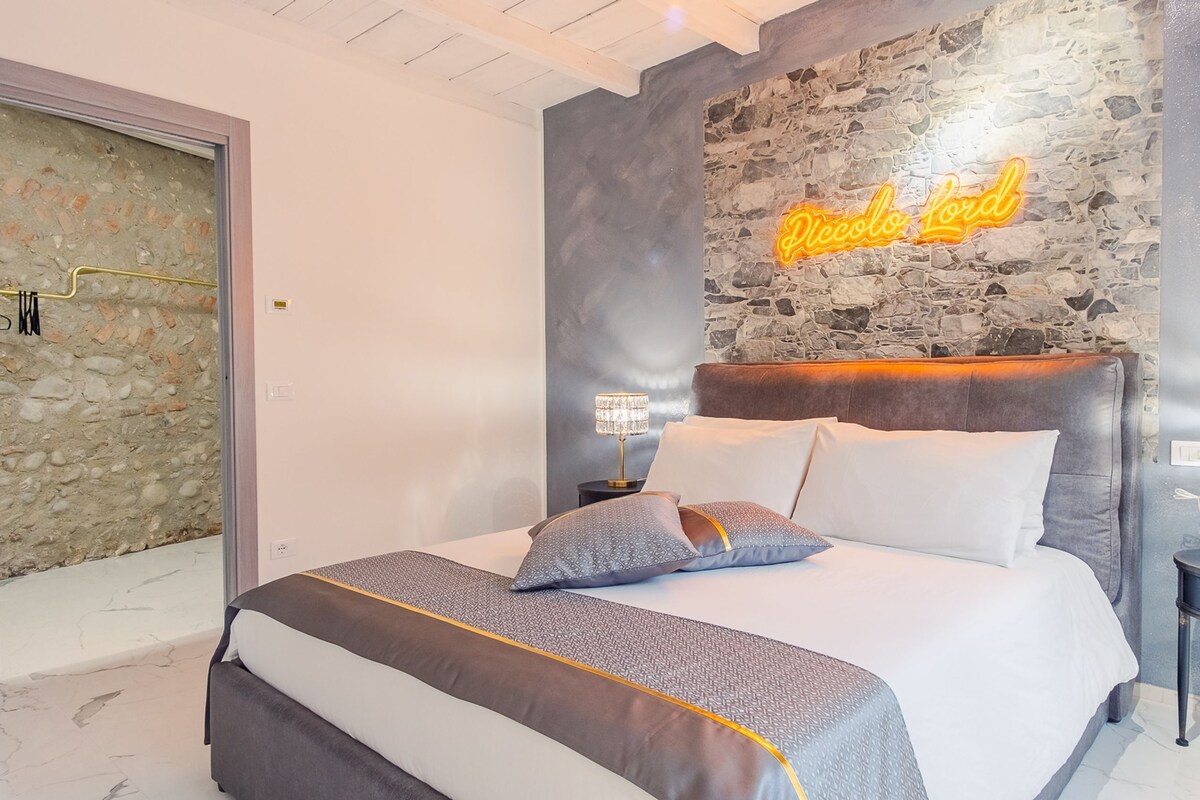 A modern bedroom with a stone accent wall, a neon sign reading "Piccolo Lord," a large bed with patterned bedding, and bedside lamps.