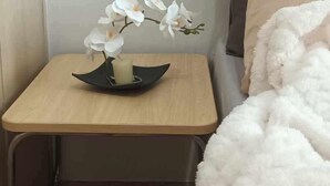 Une chambre avec une table de chevet en bois, une orchidée blanche, une bougie et une couverture blanche et moelleuse.
