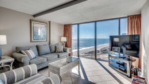 Ein modernes Wohnzimmer mit großem Fenster mit Blick auf einen Strand, einem Sofa, einem Couchtisch und einem Fernseher.