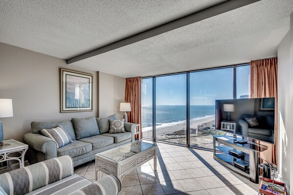 A modern living room with a large window overlooking a beach, a sofa, a coffee table, and a TV.