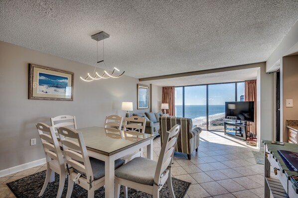 A dining area with a table and chairs, a television, and a view of the ocean.