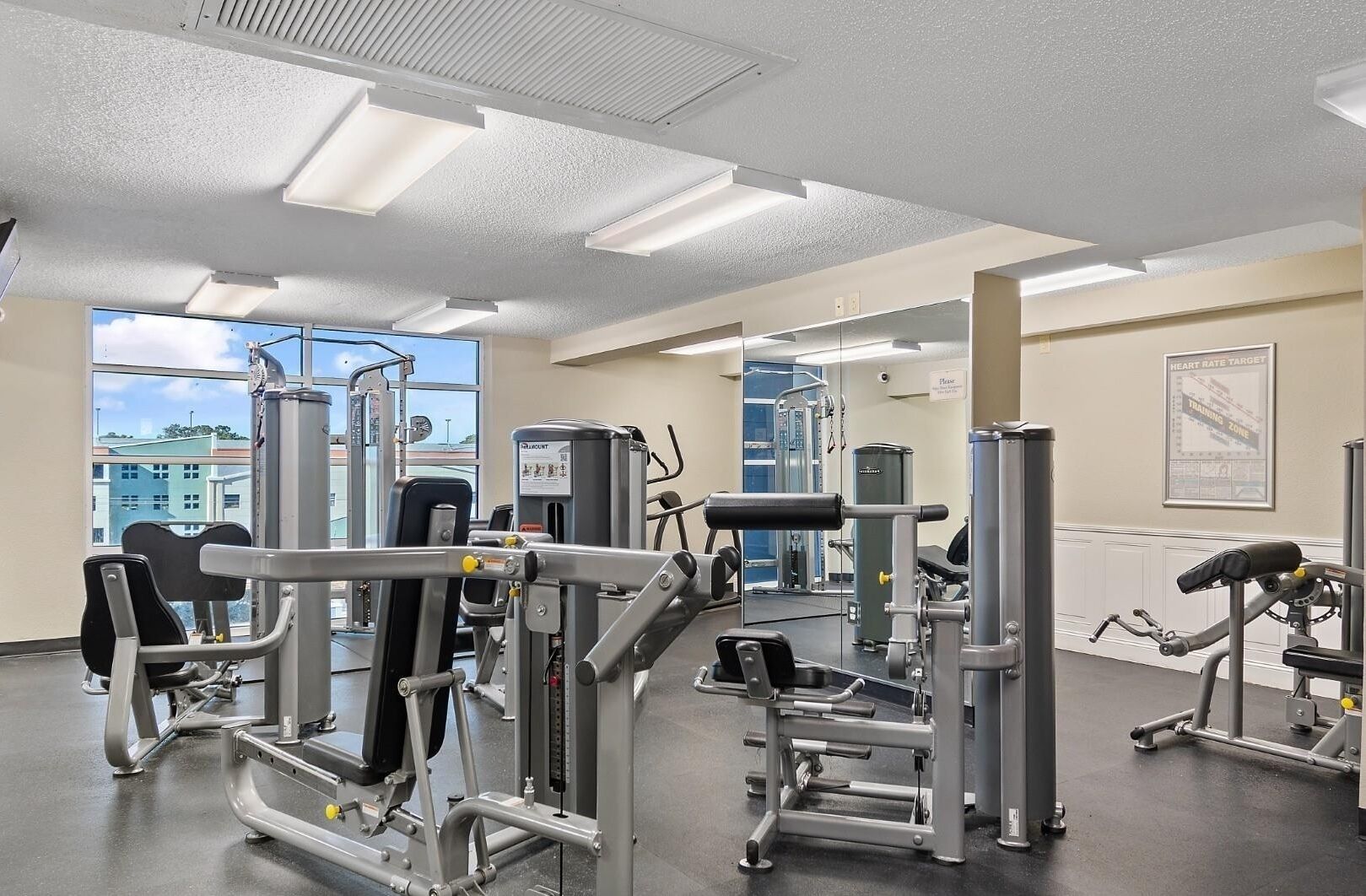 A gym with various exercise machines, large windows, and a mirror wall.