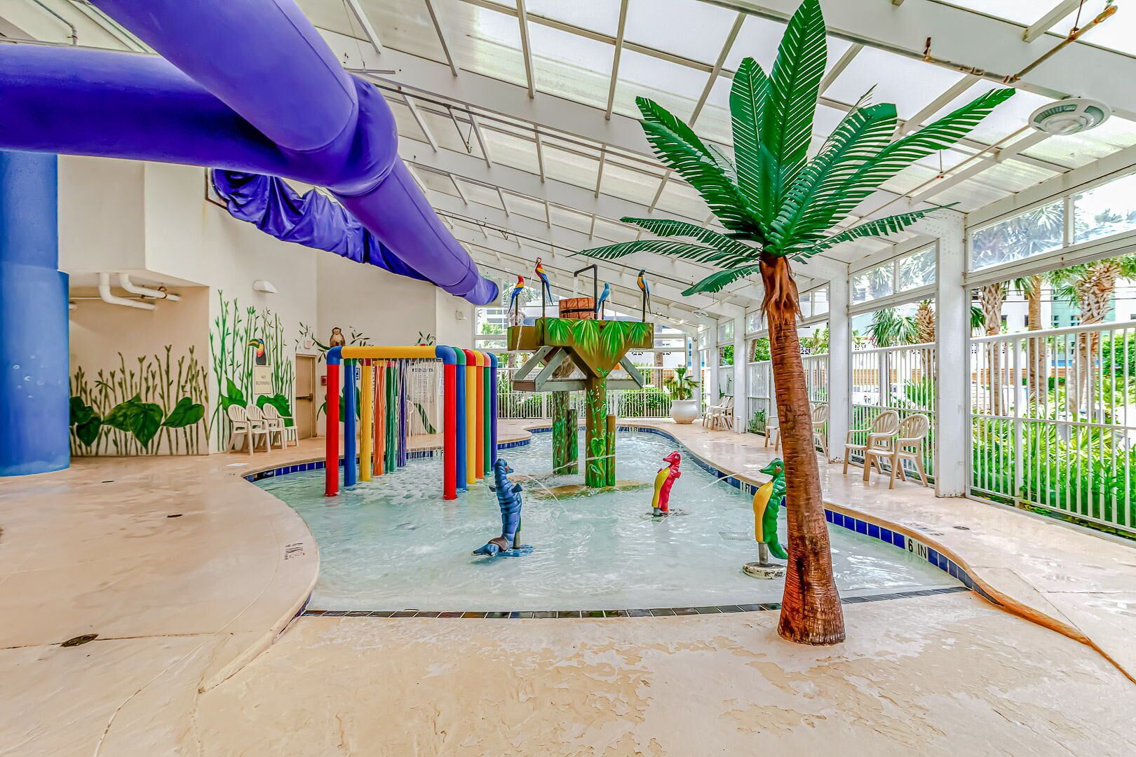 Área de recreação aquática interna com escorregadores e piscina, rodeada por palmeiras e cadeiras.