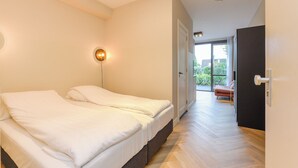 Ein Schlafzimmer mit Bett, Wandleuchte, einer Tür, die auf einen Balkon führt, und Blick ins Freie.