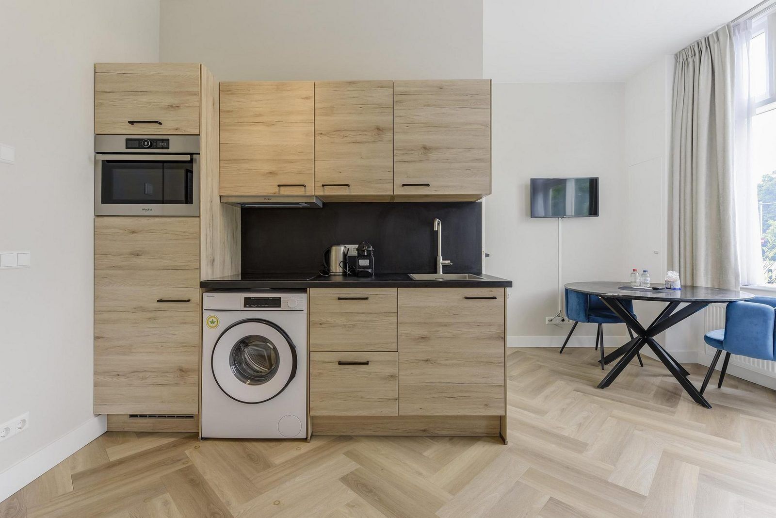 A compact kitchen with wooden cabinetry, a built-in oven, a washing machine, and a dining area with a table and chairs.