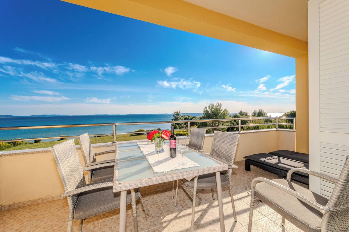 A balcony with wicker furniture, a table with a wine bottle and glasses, and a view of the sea.