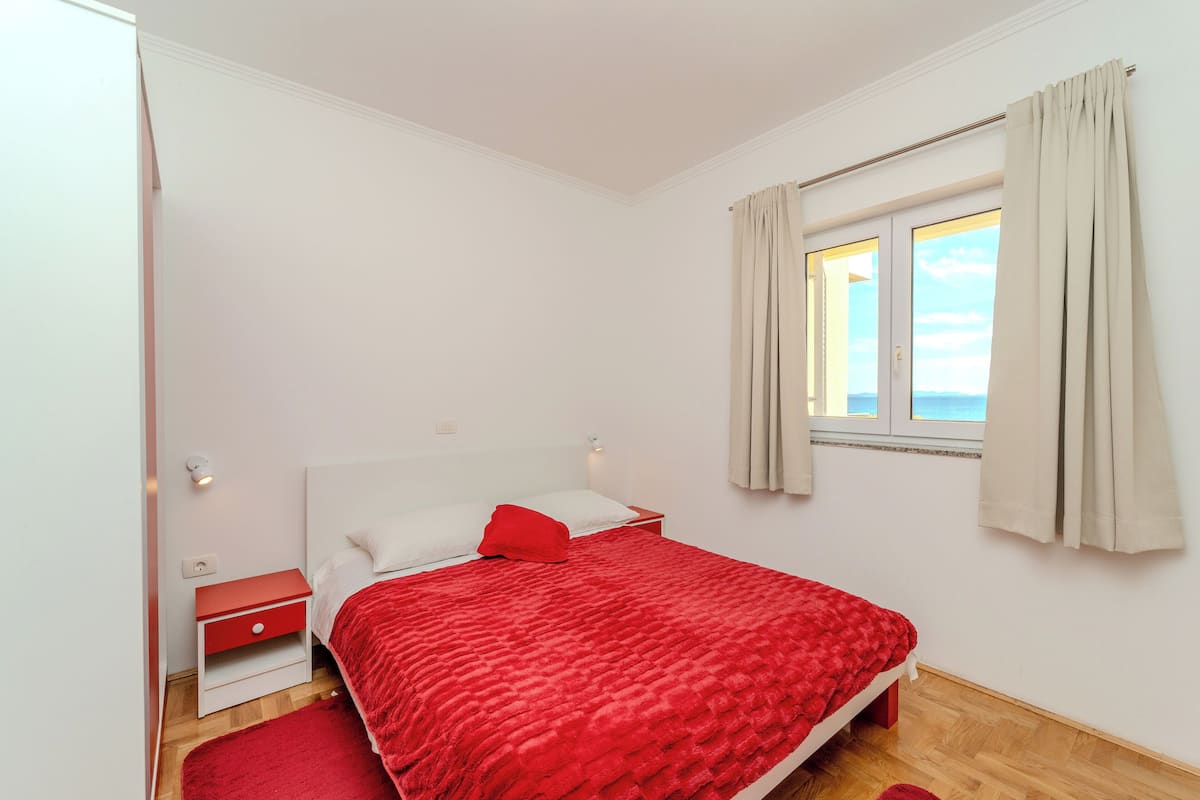 A bedroom with a bed, a nightstand, a red rug, a window with curtains, and a view of the sky.