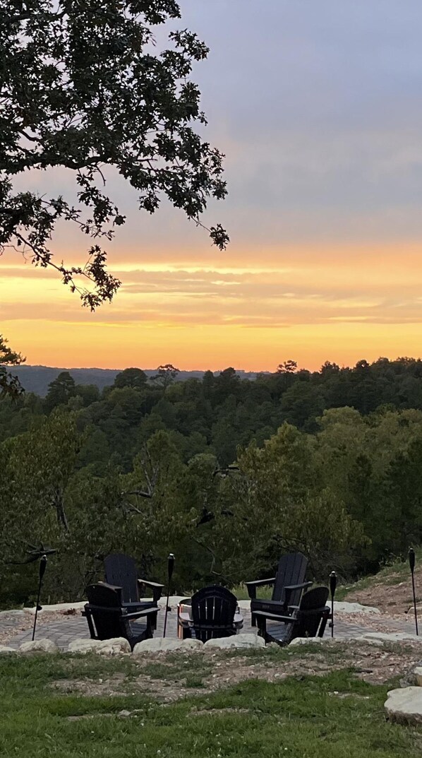 Näkymä metsäiselle alueelle auringonlaskun aikaan. Etualalla on kolme Adirondack-tuolia ja nuotiopaikka.