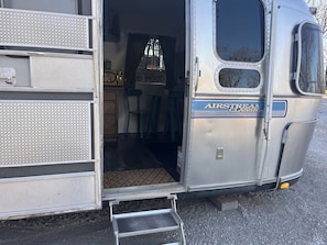 Een oude camper met een openstaande deur, waar een keuken te zien is met een fornuis, stoelen en een raam.