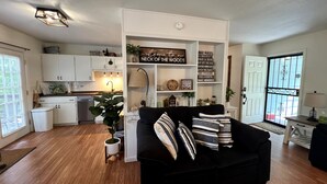 A modern living room with a black sofa, wooden flooring, a kitchen with white cabinets, and a decorative sign on the wall. - Cozy Home - Fire Pit - Seasonal Fishing (Williford)