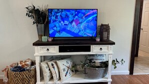 A television on a white entertainment unit with a rustic design, a small basket, and decorative pillows. - Cozy Home - Fire Pit - Seasonal Fishing (Williford)