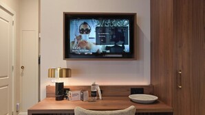 A modern hotel room with a wooden desk, a flat-screen TV mounted on the wall, a lamp, a water bottle, and a bowl. - Novallure Downtown (The Hague)