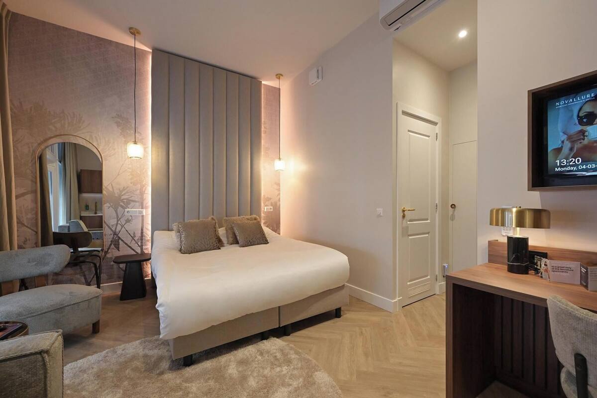 A modern hotel room with a large bed, a desk with a lamp, a TV, and a mirror.
