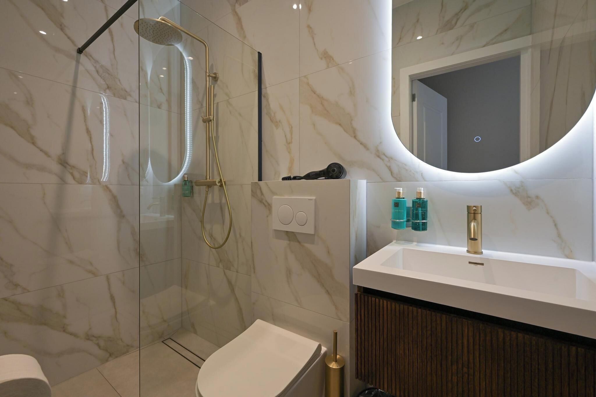 Een moderne badkamer met een marmeren vloer, een douche met glazen wanden, een witte wastafel en een houten wastafelmeubel.