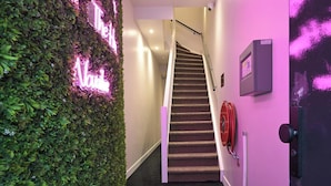 A staircase with a carpeted handrail, a fire extinguisher mounted on the wall, and a neon sign on the left. - Novallure Downtown (The Hague)