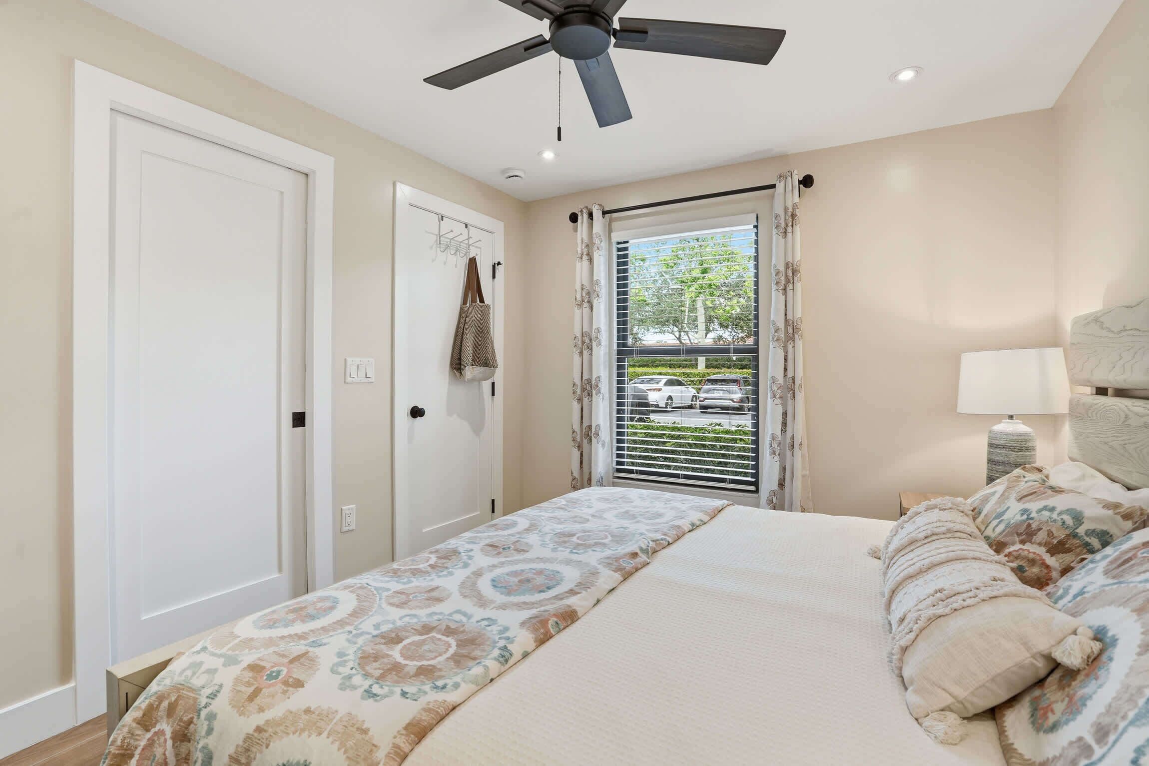A bedroom with a bed, a ceiling fan, a window with curtains, a nightstand with a lamp, and a door with a coat hanging on it.