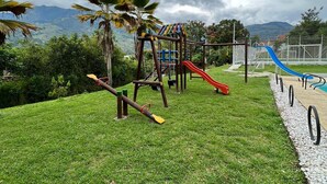 A playground with swings, slides, and climbing structures, surrounded by grass and palm trees, with mountains in the background.