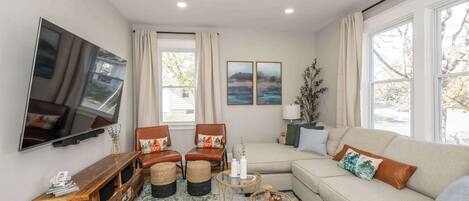 A living room with a flat-screen TV, a wooden entertainment unit, a beige sofa, two armchairs, a coffee table, and a large window with curtains.