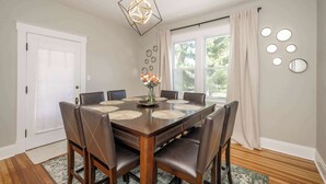 Comedor con mesa de madera, seis sillas, un florero con flores y una lámpara colgante de diseño geométrico.