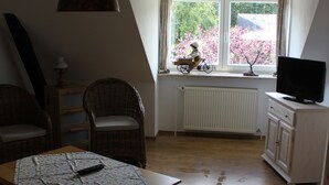 A room with a wooden table, wicker chairs, a television on a cabinet, and a window with a view of trees. - Vacation apartment Molly (Brunsbüttel)