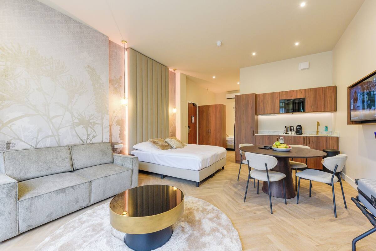Una habitación de hotel moderna con sofá, una mesa redonda, cama, mesa de comedor con sillas y zona de cocina.