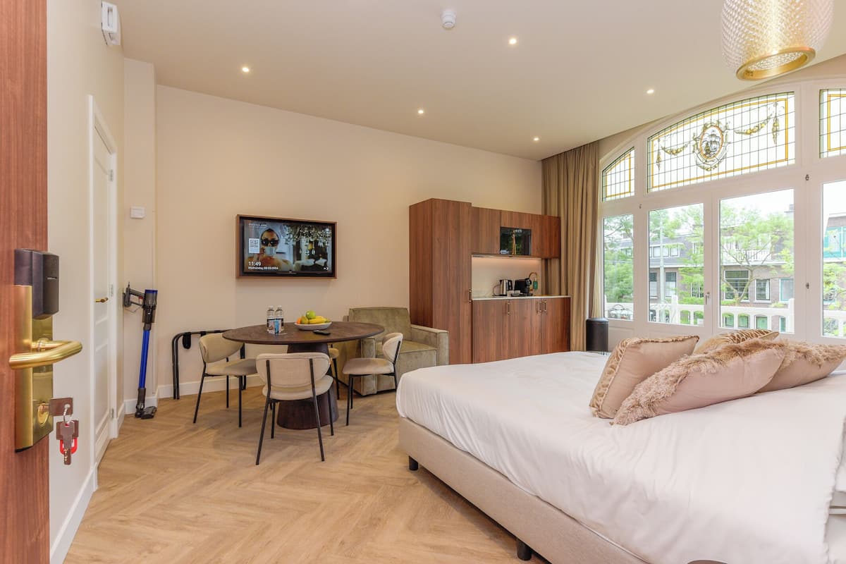 Habitación de hotel con cama, una mesa de comedor con sillas, un televisor en la pared y una ventana con cortinas.