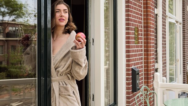 A person in a robe standing at a glass door, with a brick wall and a green metal chair visible. - Novallure Villa Margaretha (Rijswijk)