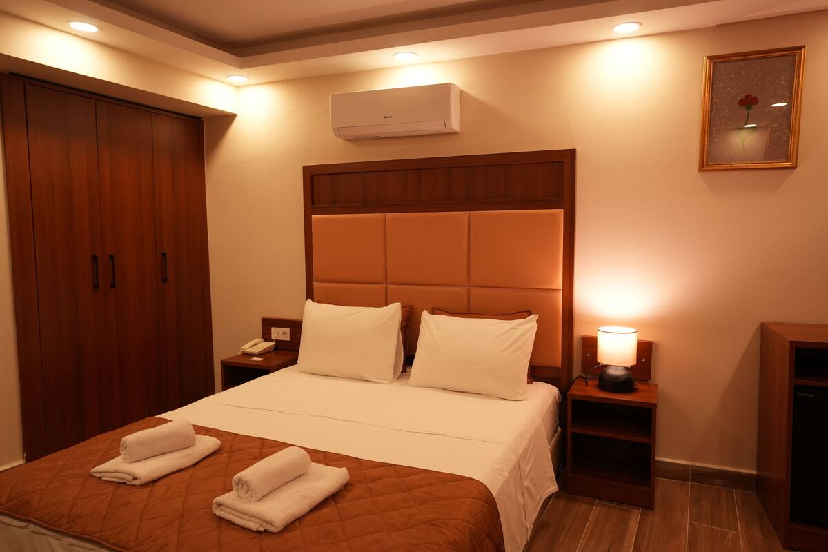 A hotel room with a large bed, wooden wardrobe, bedside tables, and a wall-mounted air conditioner.