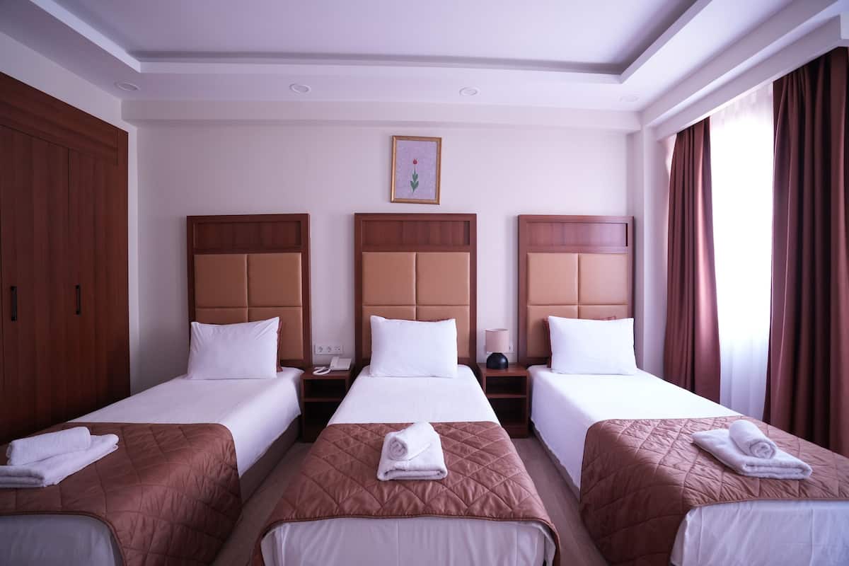 A hotel room with three single beds, each with a brown quilt and white linens. There are wooden headboards, a framed picture on the wall, and a window with curtains.