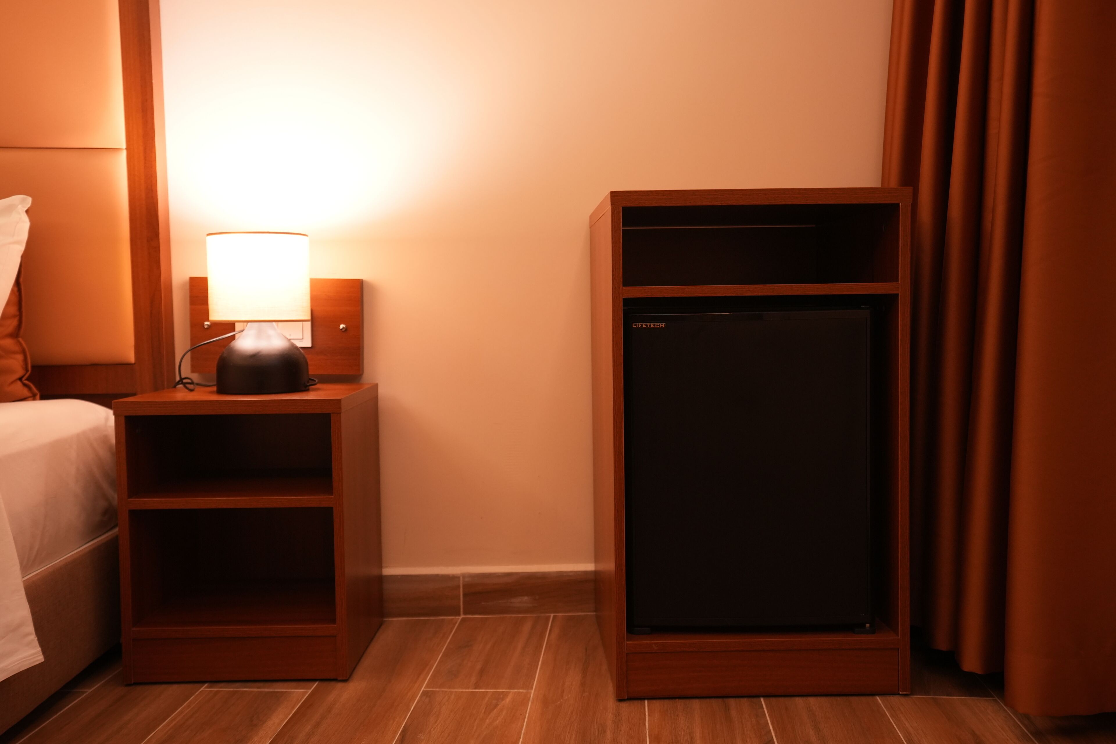 A hotel room with a bed, a nightstand with a lamp, and a minibar.