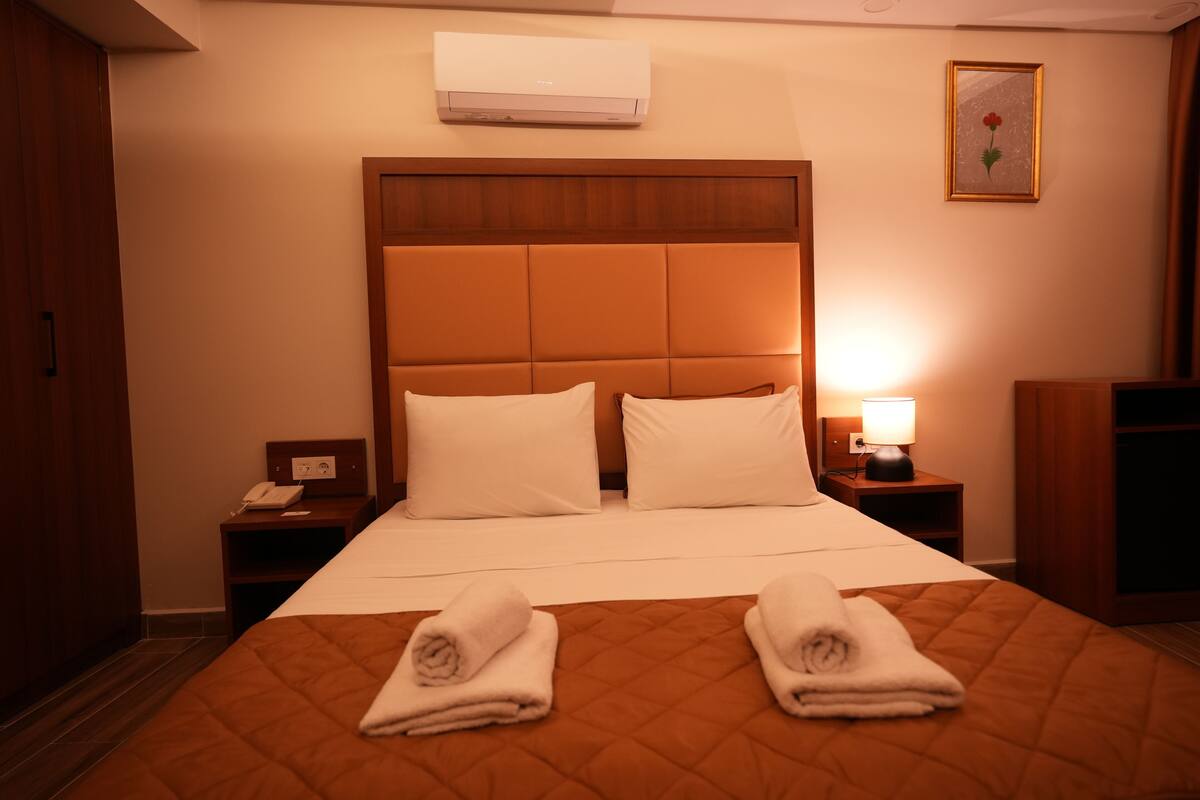 A hotel room with a large bed, two bedside tables, a nightstand, an air conditioner, and a framed picture on the wall.