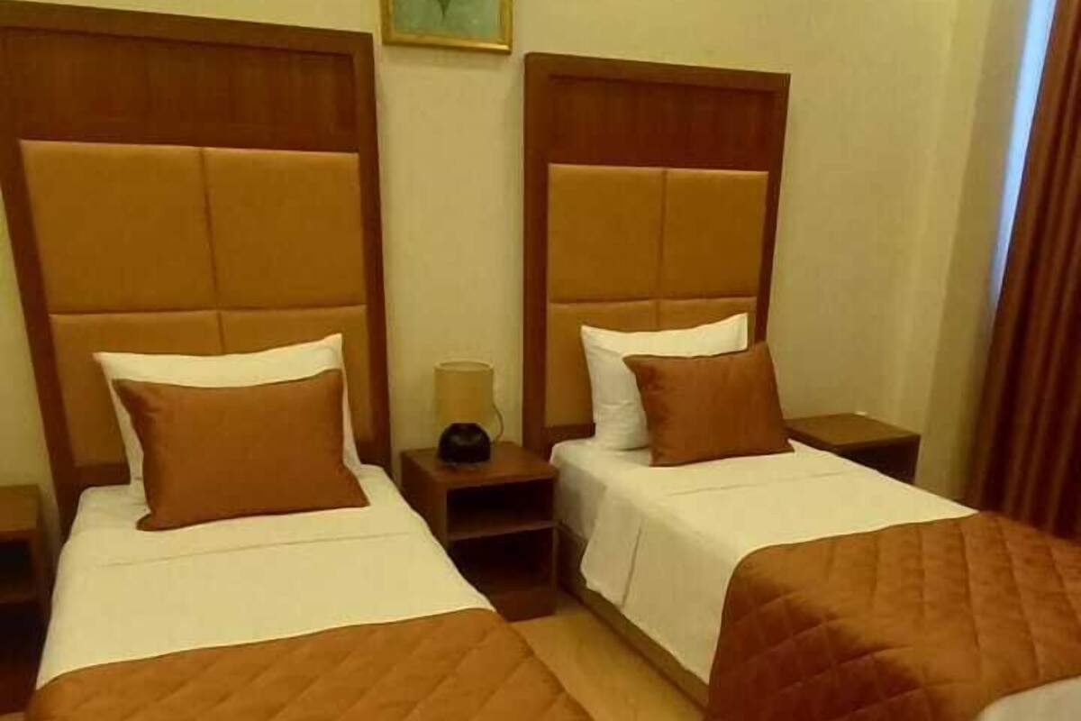 A hotel room with two beds, each with a wooden headboard and a brown quilt. There is a nightstand between the beds, and a painting on the wall.