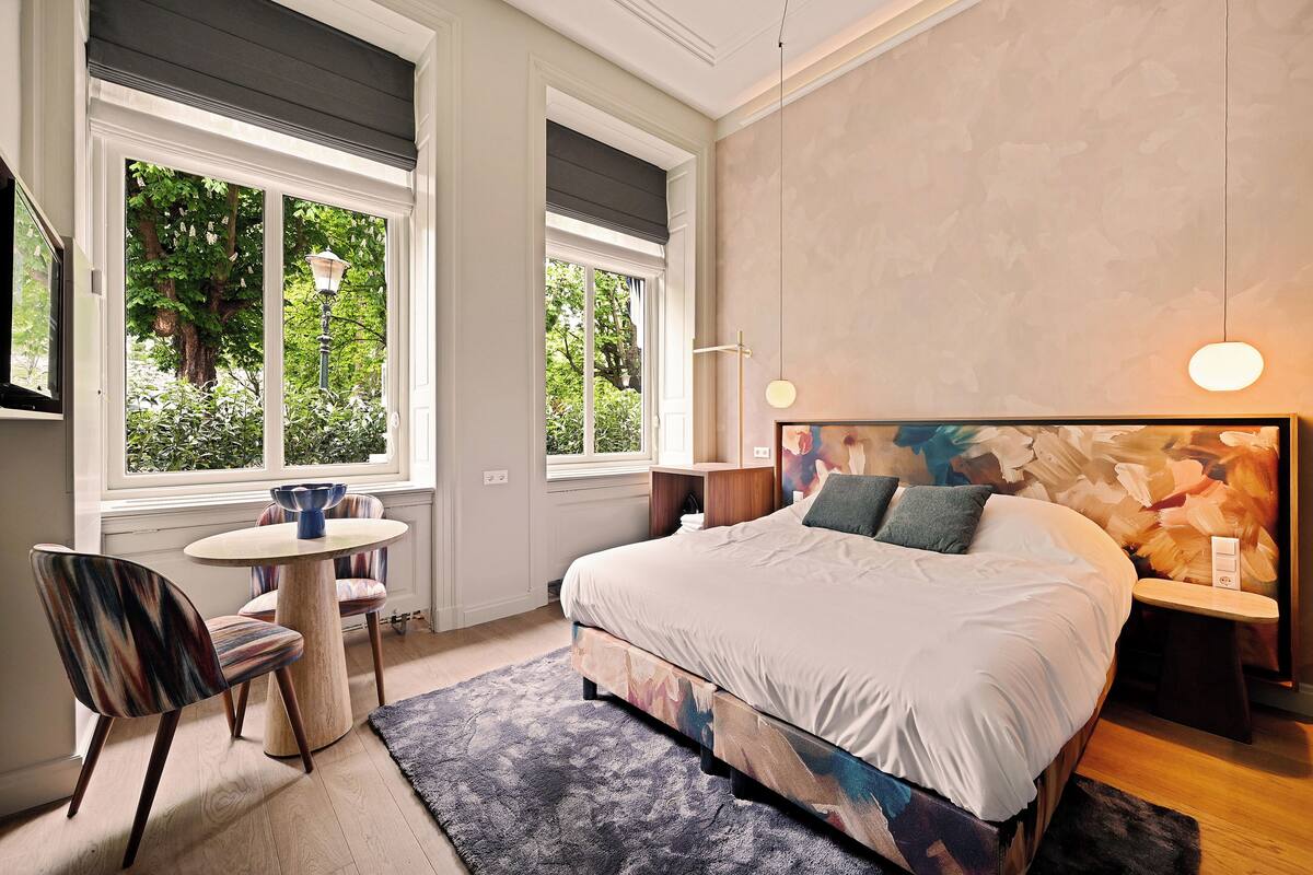 Ein Schlafzimmer mit einem großen Bett, einem runden Esstisch und zwei Fenstern mit Blick ins Grüne.