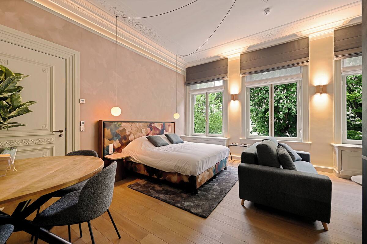 Ein Schlafzimmer mit einem großen Bett, einem Esstisch und einem Sofa. Fenster bieten Blick auf grüne Umgebung.