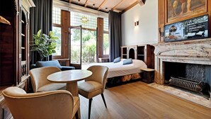 Une chambre d’hôtel comprenant un lit, deux fauteuils, une petite table, une cheminée et une grande fenêtre avec des rideaux.