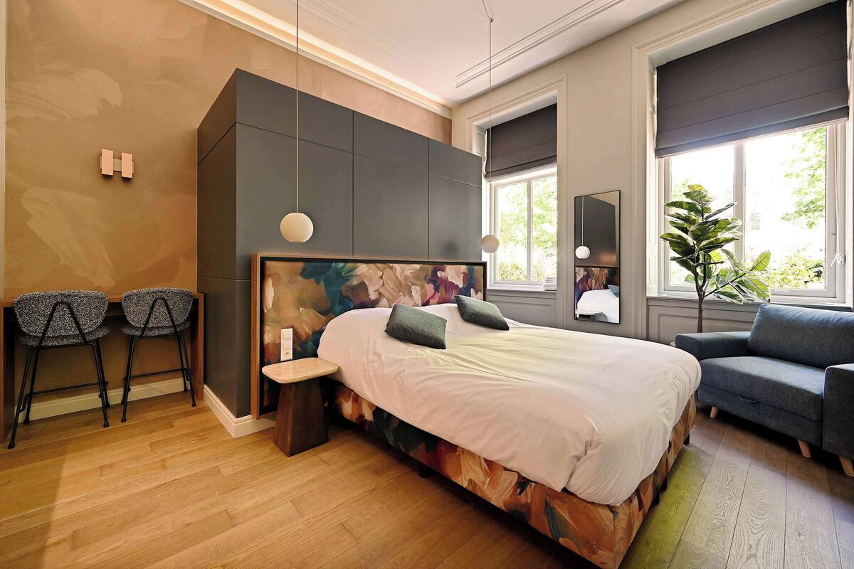 Ein modernes Schlafzimmer mit einem großen Bett, zwei Stühlen, einem Sofa und einem großen Fenster mit Blick auf Grünflächen.
