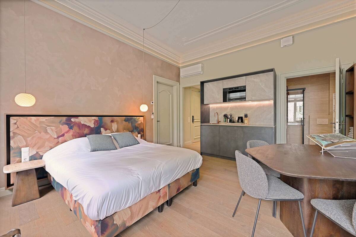 Ein Schlafzimmer mit Bett, Nachttisch und einem kleinen Schreibtisch. Es gibt eine Küchenzeile mit Mikrowelle, Herd und Schränken. Außerdem ist ein Esstisch mit Stühlen zu sehen.