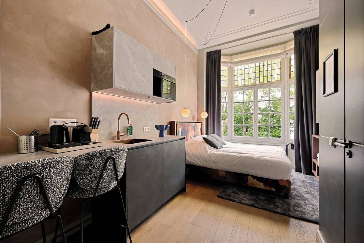 Ein kleines Hotelzimmer mit Küchenzeile, einem Bett und einem großen Fenster mit Blick auf Grünflächen.
