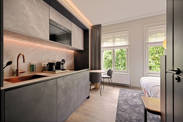 A modern kitchen with a built-in oven, a coffee maker, and a sink. There is a dining area with a round table and two chairs. The room has large windows with curtains and a bed with a gray headboard. - Novallure Nassau Residence (The Hague)