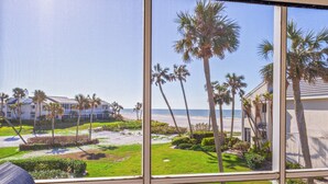 A view from a window overlooking a beach with palm trees, a grassy area, and buildings in the background. - Gulf Views & Cozy Comfort w Large Lanai Tern's Nest #22 Condo by RVA (Longboat Key)