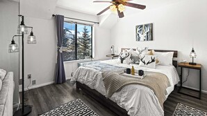 A bedroom with a large bed, a ceiling fan, a bedside table with a lamp, a window with a view of trees, and a floor lamp. - Cozy PC Condo | Top Floor Studio (Park City)