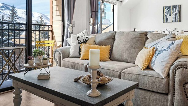 A cozy living room with a grey sofa, a white table, and a view of snow-covered mountains through large windows. - Cozy PC Condo | Top Floor Studio (Park City)