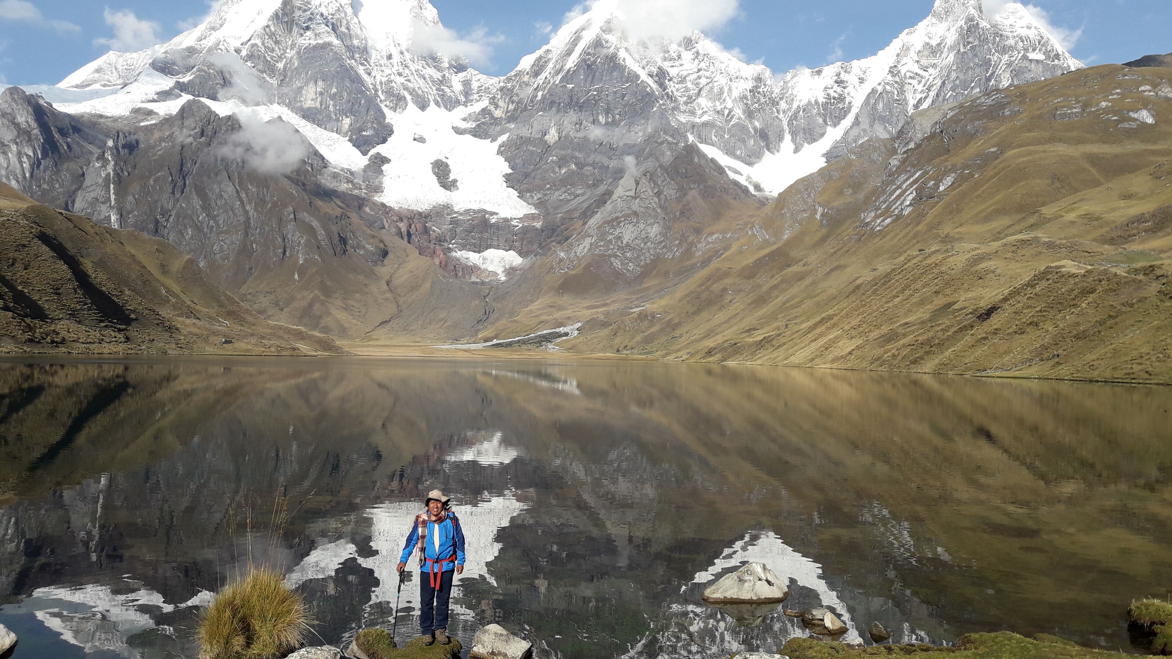Una persona de pie junto a un lago tranquilo, con montañas nevadas al fondo.
