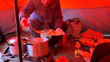 A person cooking outdoors with pots and pans on a portable stove, surrounded by various cooking utensils and food items.