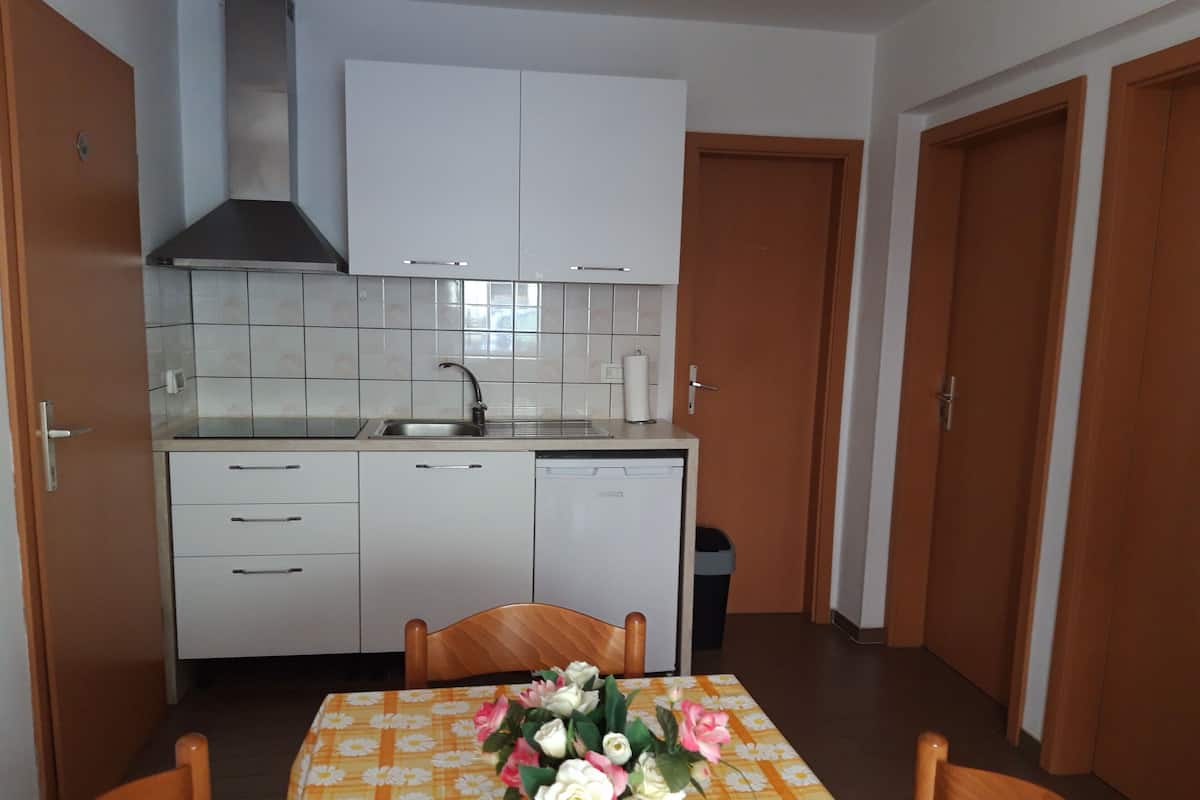 Une cuisine compacte avec des meubles blancs, un évier et un lave-vaisselle. On y trouve une table à manger avec des chaises et un vase de fleurs.