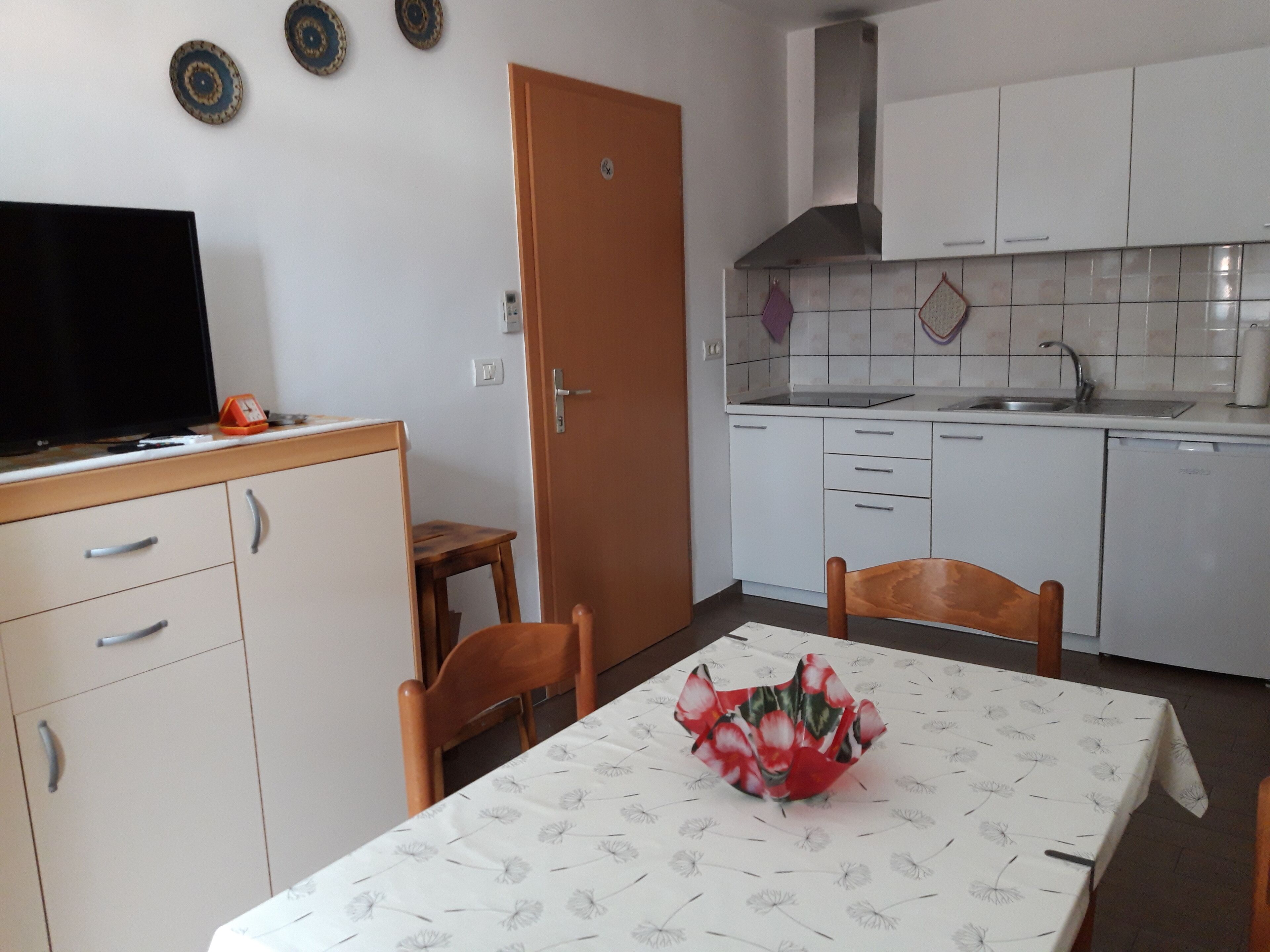 A compact kitchen with white cabinets, a built-in oven, and a sink. There's a dining table with a floral tablecloth and a vase of flowers.