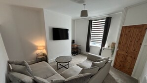 Una sala de estar con sofá, televisión, lámpara, una mesita y una ventana con cortinas.